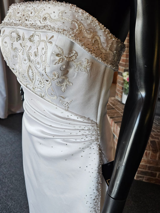 Maggie Sottero Wedding Gown  This gorgeous designer gown features a straight, banded neckline with lots of beads and embroidery. The asymmetrical side sweep adds interest and creates a lovely waistline. The creamy white satin is lustrous and falls to a small sweep train.   This gown is part of our consignment collection and has been worn before and slightly altered. It is in excellent condition and is ready to be loved again!